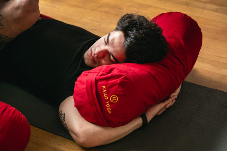 Close up of student lying down with head on bolster, hands interlocked underneath, neck turned to left