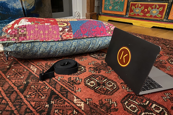 Home practice equipment of colorful pillow, black strap, and computer with Kaiut Yoga sticker on a patterned rug
